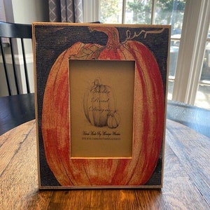 May include: A rectangular picture frame with a pumpkin design. The frame has a gold border and a cutout for a photo. The pumpkin is orange and red with a green stem, set against a dark blue background. The frame has text that reads "October Road Designs."