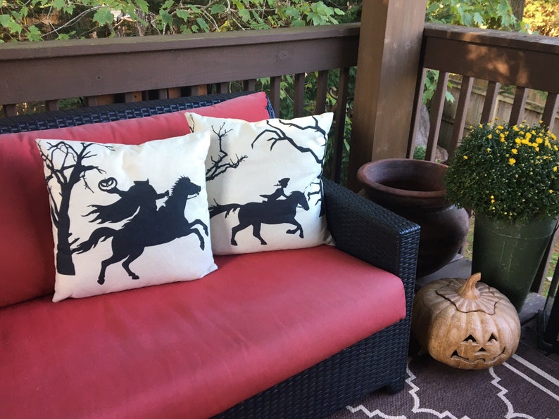 May include: Two white throw pillows with black silhouettes of a rider on a horse in a spooky forest setting. The pillows are on a red wicker couch on a porch with a carved pumpkin and potted plants.