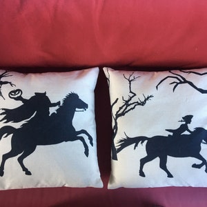 May include: Two decorative throw pillows with a white background and black silhouettes of a horse and rider. One pillow features a silhouette of a rider holding a jack-o-lantern, while the other pillow features a silhouette of a rider with a long flowing cape.