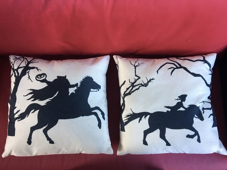 May include: Two decorative throw pillows with a white background and black silhouettes of a horse and rider. One pillow features a silhouette of a rider holding a jack-o-lantern, while the other pillow features a silhouette of a rider with a long flowing cape.