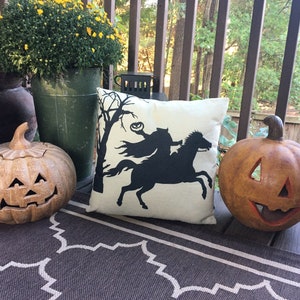 May include: Two brown ceramic jack-o'-lanterns and a white pillow with a black silhouette of a witch riding a horse on a porch with a brown rug.