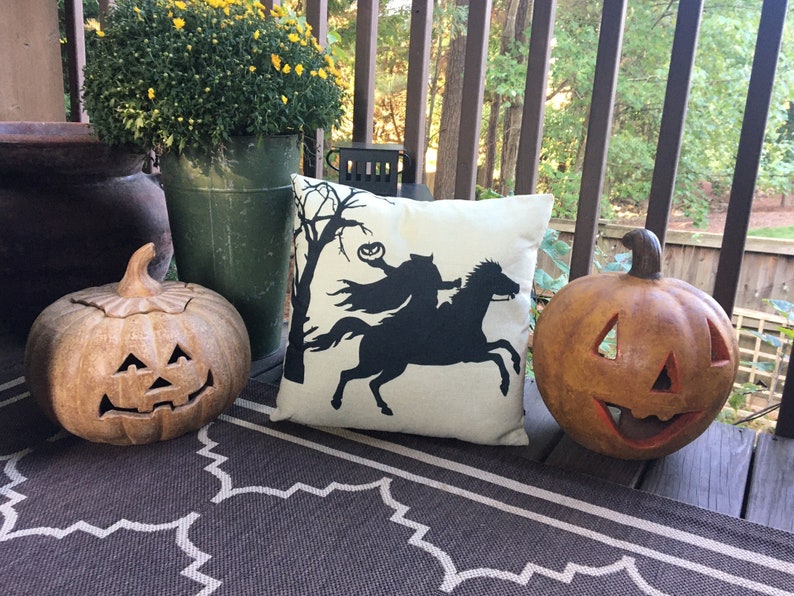 May include: Two brown ceramic jack-o'-lanterns and a white pillow with a black silhouette of a witch riding a horse on a porch with a brown rug.