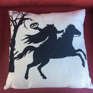 May include: A white throw pillow with a black silhouette of a rider on a horse holding a jack-o-lantern. The rider is wearing a cape and the horse is running.