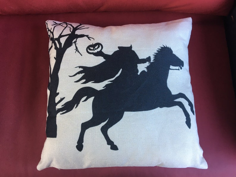 May include: A white throw pillow with a black silhouette of a rider on a horse holding a jack-o-lantern. The rider is wearing a cape and the horse is running.