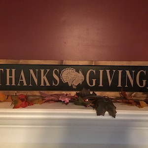 May include: A wooden sign with a black background and brown lettering that reads "THANKSGIVING". A brown silhouette of a turkey is centered between the words. The sign is framed in a rustic brown wood frame.