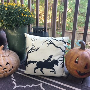 May include: A white throw pillow with a black silhouette of a woman riding a horse through a spooky forest. The pillow is on a brown and white rug on a porch with two pumpkins.