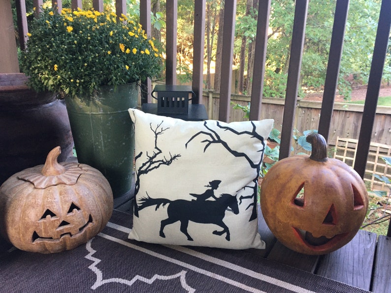 May include: A white throw pillow with a black silhouette of a woman riding a horse through a spooky forest. The pillow is on a brown and white rug on a porch with two pumpkins.