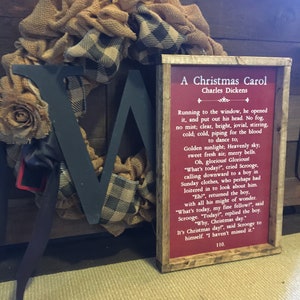 Peut inclure: Une pancarte rouge encadr&eacute;e avec une bordure blanche et le texte "A Christmas Carol" de Charles Dickens. Le texte sur la pancarte est un extrait du livre. La pancarte est appuy&eacute;e contre une couronne en toile de jute et en carreaux noirs et blancs.