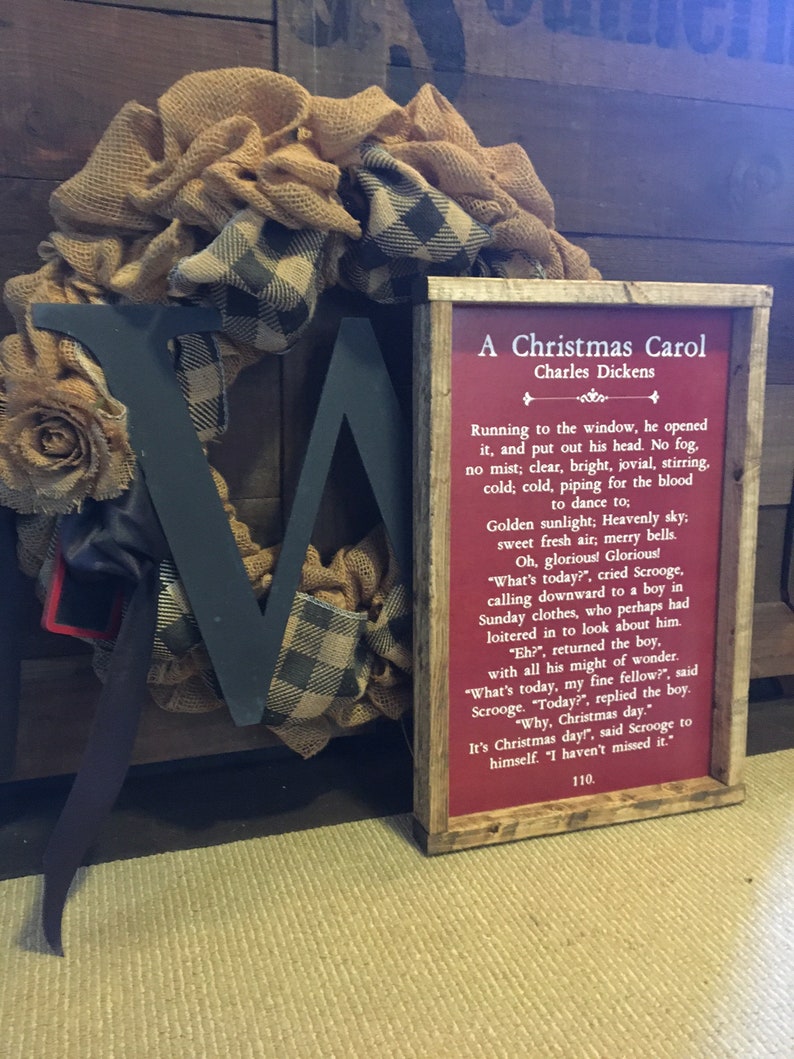 Peut inclure: Une pancarte rouge encadr&eacute;e avec une bordure blanche et le texte "A Christmas Carol" de Charles Dickens. Le texte sur la pancarte est un extrait du livre. La pancarte est appuy&eacute;e contre une couronne en toile de jute et en carreaux noirs et blancs.
