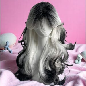 Puede incluir: Peluca larga y ondulada, bicolor. Presenta una mezcla de mechones negros y blancos, peinada con raya a un lado y flequillo. La peluca se muestra sobre una cabeza de maniquí, sobre un fondo rosa.