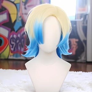 Op de afbeelding: Tweekleurige pruik met een blonde bovenkant en blauwe punten, gepresenteerd op een witte mannequinhoofd. De pruik heeft een korte, gelaagde stijl. De achtergrond heeft een kleurrijke graffiti-muurschildering en een wit oppervlak.