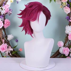 May include: A magenta wig with a layered, spiky design is presented on a white mannequin head. The wig is the central element, set against a backdrop of a floral wreath with pink and white roses. The wig's colour is a deep, rich shade.