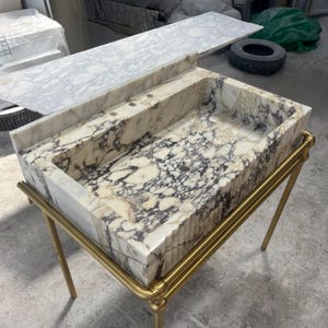 Può includere: Un lavabo rettangolare in marmo con una struttura in metallo color oro. Il lavabo presenta un motivo color crema e grigio, con due fori per i rubinetti. Un piano di lavoro in marmo abbinato poggia dietro il lavabo. È un elemento per il bagno.
