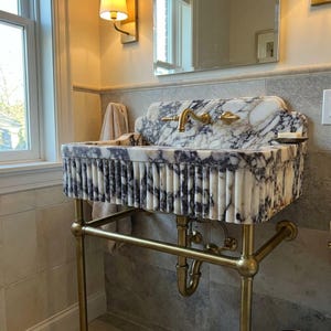 May include: A wall-mounted marble sink featuring a white and dark purple veined pattern with a fluted exterior design. The basin is supported by a brass metal frame with exposed plumbing pipes and includes a matching brass faucet fixture.