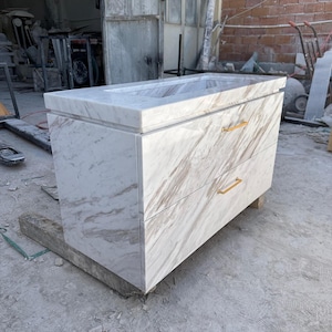 May include: A marble vanity with a rectangular sink basin and two drawers. The white marble has brown veining, and the drawers have gold-colored handles. The vanity is set on a wooden base.