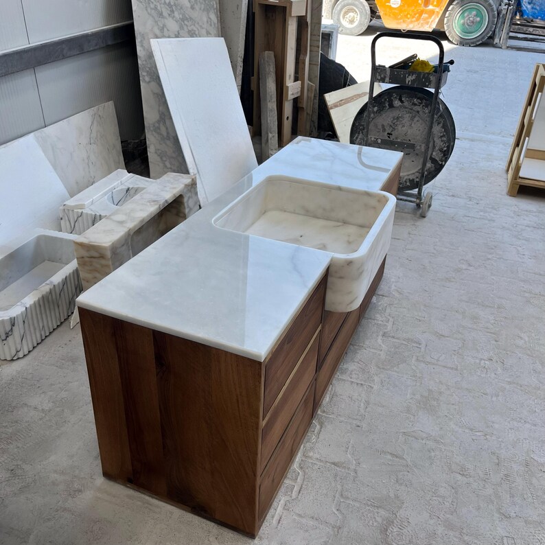 Puede incluir: Una isla de cocina hecha a medida con una encimera de m&aacute;rmol blanco y un fregadero integrado. La base est&aacute; construida con madera oscura con varios cajones. En el fondo se ven losas y fregaderos de m&aacute;rmol adicionales.