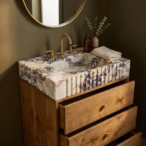 May include: A bathroom vanity featuring a white and purple marble sink with a fluted exterior design, paired with a wooden cabinet base with two drawers. A gold faucet is mounted on the sink, and a folded white cloth rests on the edge.