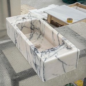 May include: A rectangular white marble sink with black veining. The sink has a rectangular basin with two faucet holes and a drain hole. The sink is made of polished marble and is suitable for a bathroom.