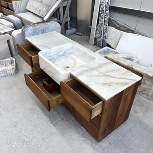 Puede incluir: Un mueble de ba&ntilde;o a medida con encimera de m&aacute;rmol y lavabo integrado. El mueble tiene una base de madera marr&oacute;n claro con varios cajones. El m&aacute;rmol presenta un veteado blanco y gris.