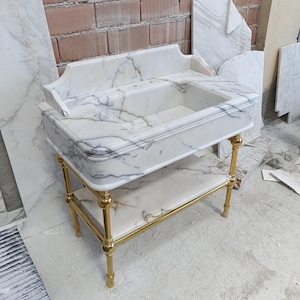 Peut inclure: Un meuble-lavabo en marbre blanc veiné de gris, avec un lavabo rectangulaire et une étagère inférieure. Le meuble est soutenu par des pieds et un cadre en métal doré ornementé. Le design est classique et élégant.