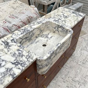 K&ouml;nnte beinhalten: Ein Sp&uuml;lbecken im Landhausstil, integriert in eine wei&szlig;-graue Marmorarbeitsplatte mit ausgepr&auml;gter Maserung. Das Becken ist auf einem dunklen Holzschrank montiert, der mehrere Schubladen mit kleinen, runden, goldfarbenen Metallkn&ouml;pfen besitzt.