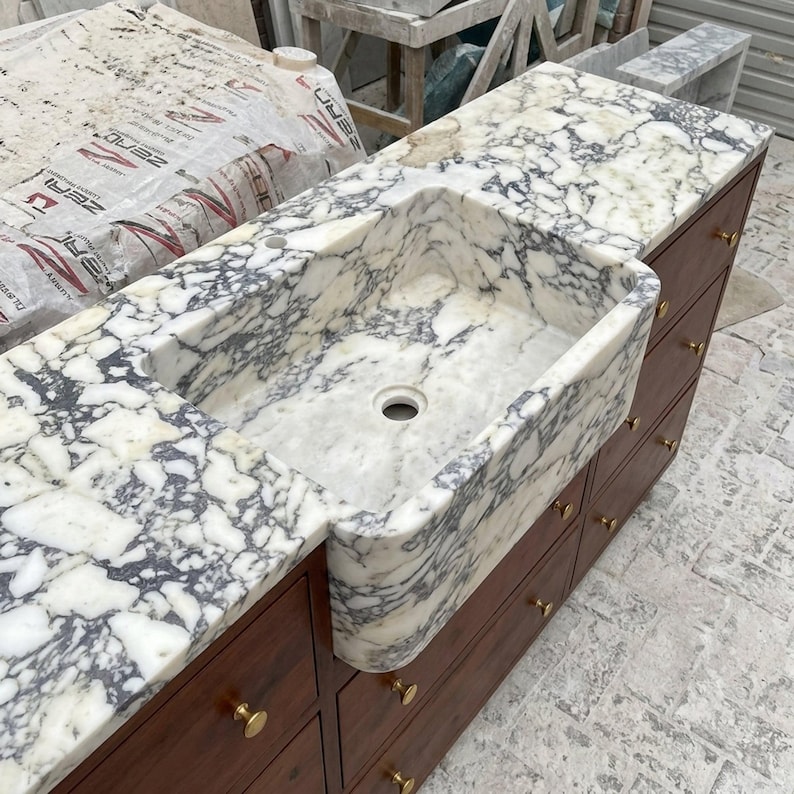 K&ouml;nnte beinhalten: Ein Sp&uuml;lbecken im Landhausstil, integriert in eine wei&szlig;-graue Marmorarbeitsplatte mit ausgepr&auml;gter Maserung. Das Becken ist auf einem dunklen Holzschrank montiert, der mehrere Schubladen mit kleinen, runden, goldfarbenen Metallkn&ouml;pfen besitzt.