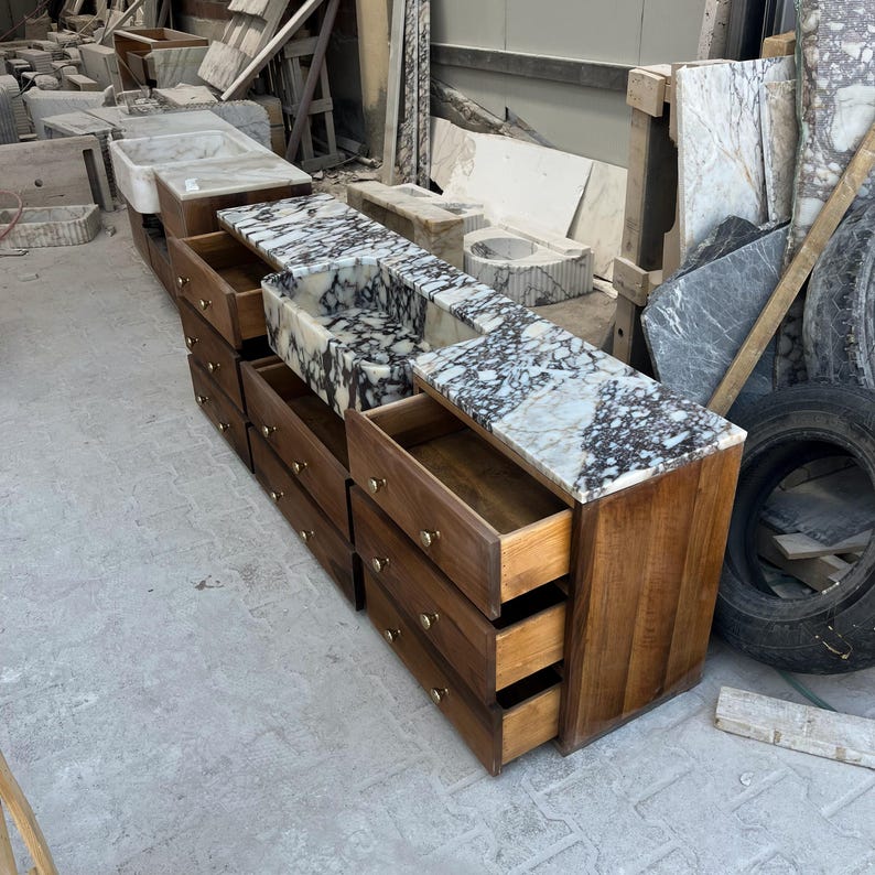 K&ouml;nnte beinhalten: Ein Waschtischunterschrank aus Holz mit einer wei&szlig;-schwarzen Marmorplatte und einem integrierten rechteckigen Waschbecken. Der Schrank verf&uuml;gt &uuml;ber drei &uuml;bereinanderliegende Schubladen mit kleinen runden messingfarbenen Kn&ouml;pfen.