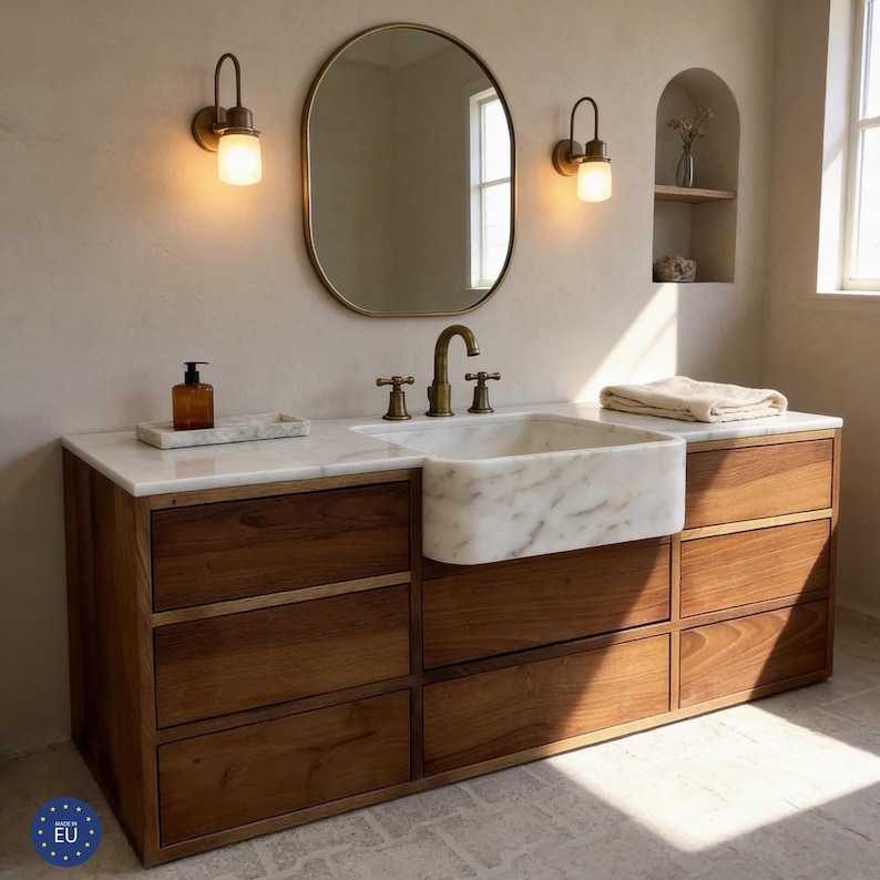 Puede incluir: Un mueble de ba&ntilde;o con encimera de m&aacute;rmol y lavabo de m&aacute;rmol blanco. El mueble tiene una base de madera con varios cajones. Un espejo ovalado con marco dorado cuelga sobre el lavabo, flanqueado por dos apliques.