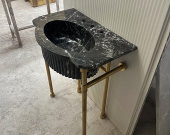 May include: A black marble vanity with white veining, featuring a built-in oval sink. The vanity has a fluted front and is supported by brass-colored metal legs. The design is a blend of classic and modern styles, suitable for a bathroom.