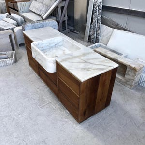 Puede incluir: Un mueble de ba&ntilde;o hecho a medida con encimera de m&aacute;rmol blanco y lavabo integrado. El mueble tiene un mueble de madera de tonos c&aacute;lidos con m&uacute;ltiples cajones. El m&aacute;rmol tiene vetas grises y marrones. El dise&ntilde;o es moderno y elegante.