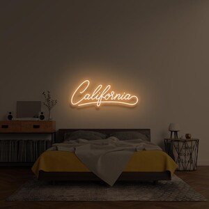 Op de afbeelding: Een oranje neonbord met het woord "California" in cursief schrift, boven een bed met gele beddengoed. De kamer heeft een modern, minimalistisch ontwerp, met een nachtkastje en een houten kast.