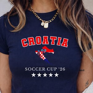 Front view of a navy blue Croatia "Soccer Cup '26" baby tee on a model. The white stars and text pop sharply against the dark fabric in a bright indoor setting.