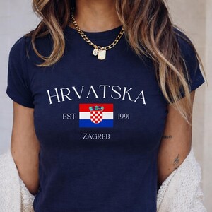 A woman modeling a deep navy blue cropped t-shirt with the contrast white "HRVATSKA ZAGREB" text and red-and-white checkered Croatian crest. The background features clean, neutral-toned modern tiles.