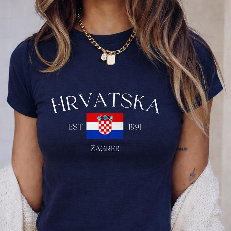 A woman modeling a deep navy blue cropped t-shirt with the contrast white "HRVATSKA ZAGREB" text and red-and-white checkered Croatian crest. The background features clean, neutral-toned modern tiles.
