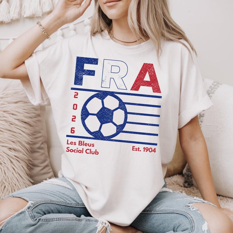 Close-up of a female model sitting on a cozy beige sofa, wearing a tucked-in ivory cream t-shirt with a blue and red distressed retro France soccer graphic, paired with ripped denim jeans.
