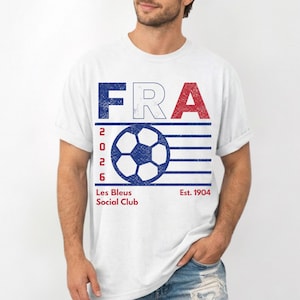 Peut inclure: Un t-shirt blanc &agrave; manches courtes avec un graphisme vieilli affichant le texte FRA en bleu, blanc et rouge. En dessous, un ballon de football bleu est entour&eacute; de lignes horizontales avec le texte vertical 2026. Inscription Les Bleus Social Club Est. 1904.