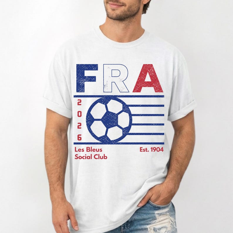 Peut inclure: Un t-shirt blanc &agrave; manches courtes avec un graphisme vieilli affichant le texte FRA en bleu, blanc et rouge. En dessous, un ballon de football bleu est entour&eacute; de lignes horizontales avec le texte vertical 2026. Inscription Les Bleus Social Club Est. 1904.