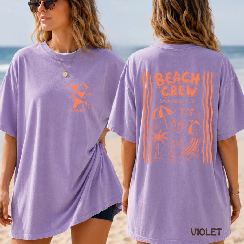 May include: A light purple short-sleeve t-shirt featuring a small orange bikini graphic on the front left chest. The back displays the text BEACH CREW, Destin Beach, FL '24, with orange illustrations of a beach umbrella, palm tree, and chair.