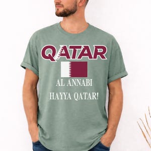 Moss T-shirt showing a vibrant Qatar soccer flag graphic and Hayya Qatar chant print.