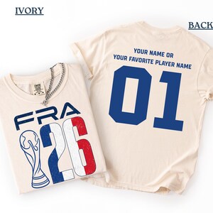 Puede incluir: Camisetas color marfil con dise&ntilde;os franceses. Una muestra "FRA 26" con un trofeo y los colores de la bandera francesa. La otra tiene un gran "01" azul en la espalda, con el texto "YOUR NAME OR YOUR FAVORITE PLAYER NAME."