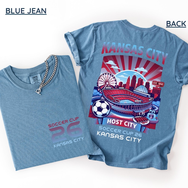 Flat lay of a "Blue Jean" colored shirt showing the front "26" logo and the detailed "Host City" back print with a silver chain accessory.