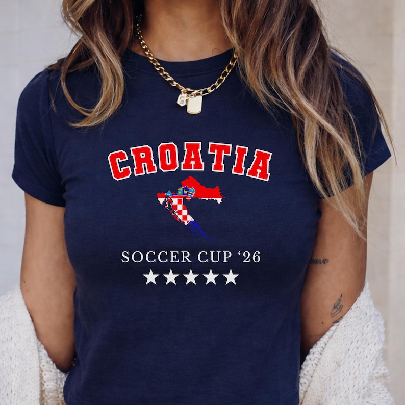Front view of a navy blue Croatia "Soccer Cup '26" baby tee on a model. The white stars and text pop sharply against the dark fabric in a bright indoor setting.