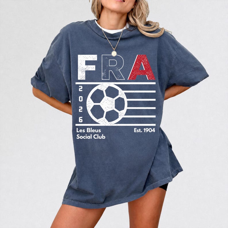 A blonde model wearing an oversized, heavy-blend pepper gray t-shirt featuring a distressed white and red "FRA 2026 Les Bleus Social Club" soccer graphic with a central football icon, shot in bright lifestyle studio lighting.