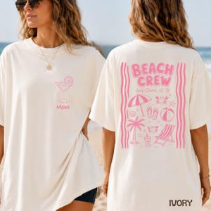 May include: An ivory t-shirt featuring a pink graphic of a cocktail glass and the word MOM on the front left chest. The back displays a large pink graphic with the text BEACH CREW, Gulf Shores, AL '26, surrounded by beach icons like a palm tree.