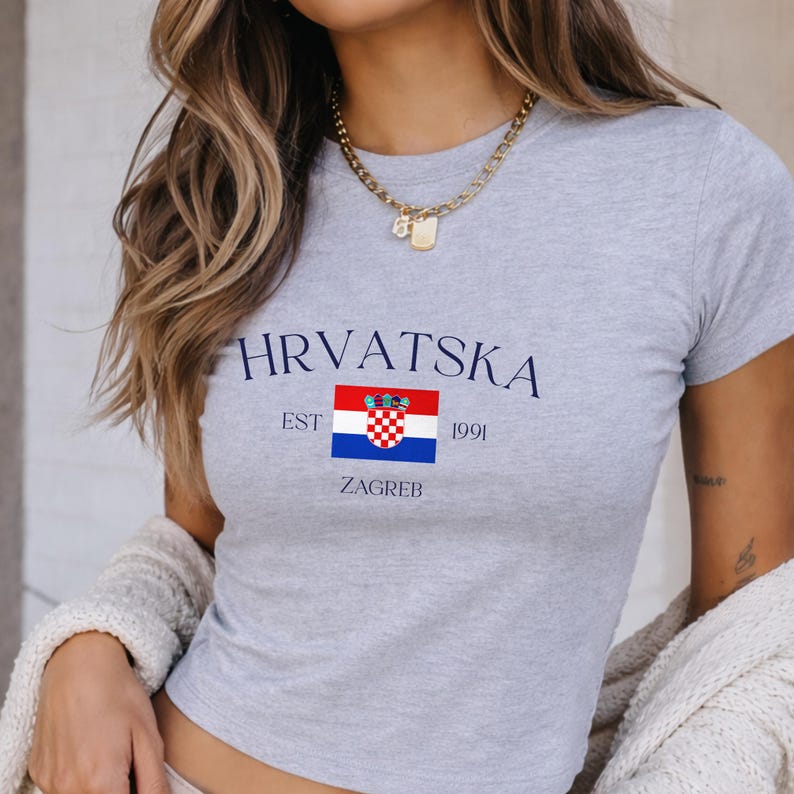 May include: A gray crop top with the word "HRVATSKA" in navy blue, along with the Croatian flag and the text "EST 1991 ZAGREB." The shirt has short sleeves and a crew neck.