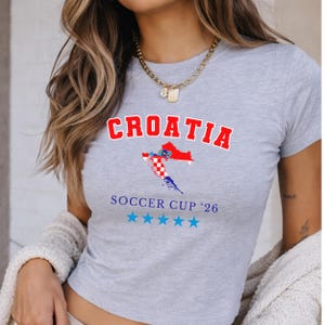 Close-up of a model wearing a ash Croatia baby tee with a gold chain and cream knit cardigan. The collegiate "CROATIA" text and flag map are centered in soft daylight.