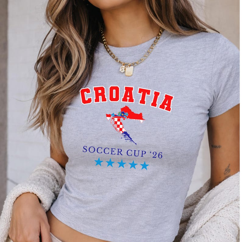 Close-up of a model wearing a ash Croatia baby tee with a gold chain and cream knit cardigan. The collegiate "CROATIA" text and flag map are centered in soft daylight.