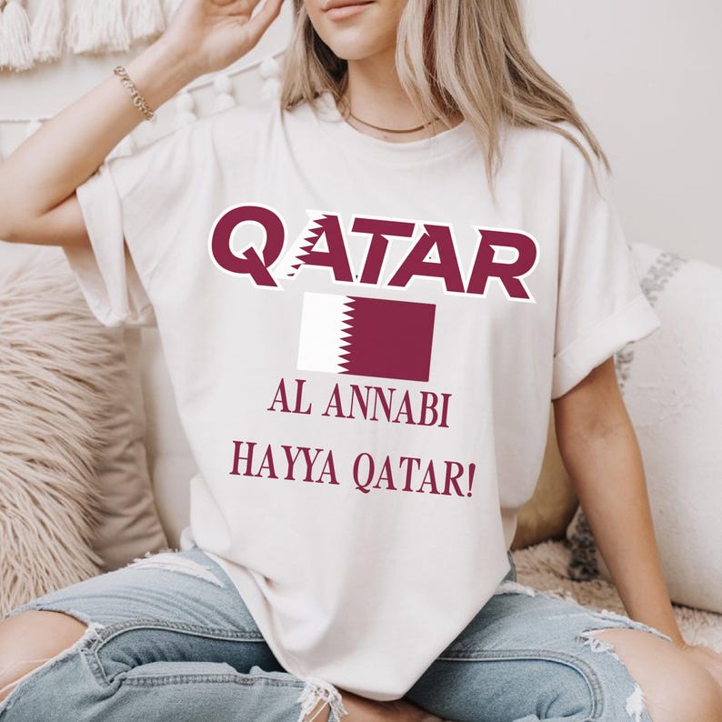 White T-shirt with dynamic Qatar text, national flag, and Hayya Qatar football supporter graphic.