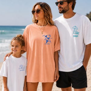 A family of three stands on a sunny sandy beach with ocean waves in the background. A man wears a white oversized tee with a blue beach chair doodle and 'DAD' text. A woman wears an orange garment with a bikini doodle and 'ASHLEY' text. A young girl smiles wearing a white tee with a blue sandcastle doodle and 'JENNIFER' text.
