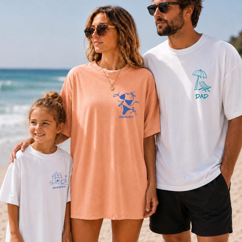 A family of three stands on a sunny sandy beach with ocean waves in the background. A man wears a white oversized tee with a blue beach chair doodle and 'DAD' text. A woman wears an orange garment with a bikini doodle and 'ASHLEY' text. A young girl smiles wearing a white tee with a blue sandcastle doodle and 'JENNIFER' text.